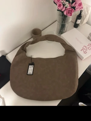 Beige axelväska i tyg - Säljer en stilren beige axelväska i mjukt tygmaterial. Väskan har en rundad form och bred axelrem som gör den bekväm att bära. Perfekt för dig som vill ha en enkel men trendig accessoar till din outfit.