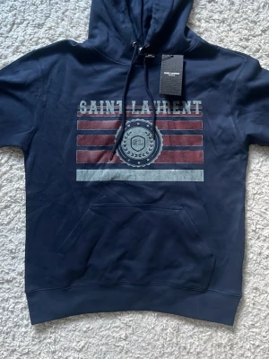 Mörkblå Saint Laurent hoodie - Snygg mörkblå hoodie från Saint Laurent med tryck framtill i rött, vitt och grått. Klassisk huva. Materialet känns mjukt och skönt, perfekt för en avslappnad stil. Saint Laurent-logga och mönster på bröstet.