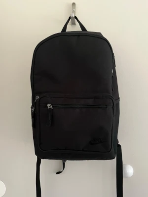Svart ryggsäck från Nike - Snygg svart ryggsäck från Nike med minimalistisk design. Ryggsäcken har ett stort huvudfack, en framficka med dragkedja och diskret Nike-logga. Tillverkad i slitstarkt tyg med justerbara axelband och bärhandtag upptill. Perfekt för skola eller cityhäng.