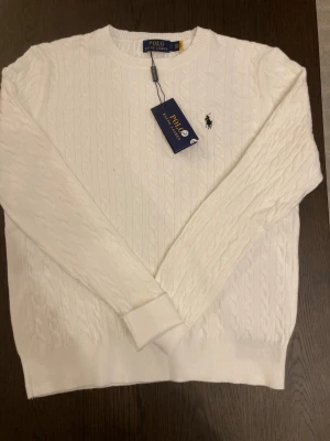 Vit kabelstickad tröja Polo Ralph Lauren - Vit kabelstickad tröja från Polo Ralph Lauren med rund halsringning och den klassiska broderade loggan i svart på bröstet. Tröjan har långa ärmar och ribbade muddar vid ärmslut och nederkant. Perfekt för en clean och stilren look. Väldigt bra skick och är i storlek L.