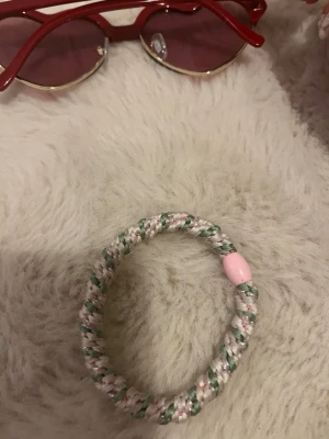 Flätad hårsnodd med pärla - Säljer en flätad hårsnodd i ljusrosa, vitt och grönt med en rosa pärldetalj. Perfekt för att piffa till din frisyr och hålla håret på plats. Tillverkad i elastiskt material med en söt och trendig look.