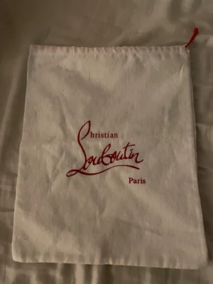 Christian Louboutin dustbag tygpåse - Beige tygpåse från Christian Louboutin med röd logotyp och dragsko upptill. Perfekt för att förvara skor eller accessoarer. Klassisk design med Paris-tryck, tillverkad i bomullstyg.