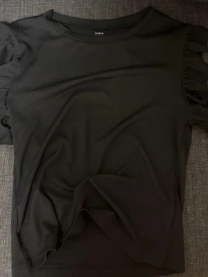 Svart ribbad topp från SHEIN - Svart ribbad topp från SHEIN med korta puffärmar och rund halsringning. Toppens material är stretchigt och sitter snyggt på kroppen. Perfekt att styla med jeans eller kjol för en trendig look.