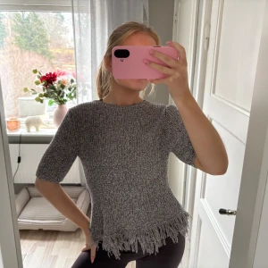 Grå stickad topp med fransar - Säljer en grå stickad topp med korta ärmar och coola fransar längs nederkanten. Tröjan har en rund halsringning och är gjord i ett mjukt stickat material som känns skönt mot huden. Perfekt för dig som gillar en unik och trendig look.