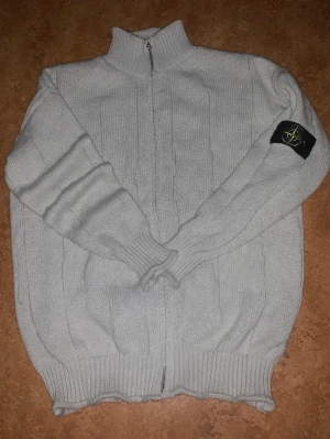 Ljusgrå stickad ziptröja Stone Island - Ljusgrå stickad ziptröja från Stone Island med hög krage och ribbade muddar. Tröjan har dragkedja framtill och den klassiska svarta loggan med kompass på vänster ärm.