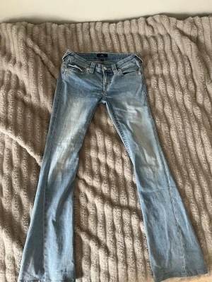 Ljusblå bootcut jeans från True Religion - Säljer ett par ljusblå bootcut jeans från True Religion med klassiska fickor och snygga slitningar framtill. Jeansen har låg midja, utsvängda ben och detaljerade bakfickor med knapp. Perfekta för dig som gillar en retro vibe. Benen är 78 cm långa från grenen och midjan i storlek w25. Knapp locket på ena fickan är sönder men fickan går att stänga. Säljer jeansen för att jag har vuxit ur dem.