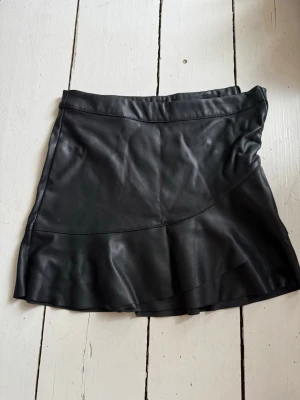 Svart kort kjol med volang - Säljer en svart kort kjol med volangdetalj längst ner. Kjolen har en snygg och modern design med skinnliknande material som ger en cool look. 