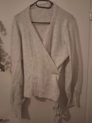 Ljusgrå omlottkofta i stickat - Mysig ljusgrå omlottkofta i stickat material med bred ribbad mudd vid ärmslut och midja. Koftan har långa ärmar och knyts i sidan för en snygg passform. Perfekt för lager på lager och enkel att styla till olika outfits.