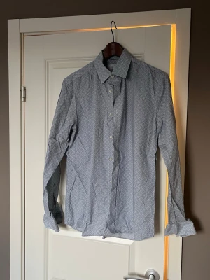 Mönstrad skjorta från Bläck - Snygg långärmad skjorta från Bläck med diskret blått mönster på vit botten. Skjortan har klassisk krage, knappar framtill och en tailored fit som ger en smalare siluett. Perfekt för dig som gillar stilrena detaljer.