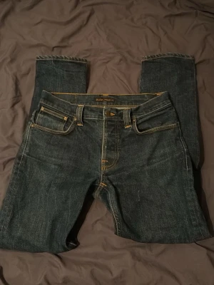 Grim tim jeans från Nudie Jeans - Säljer ett par mörkblå jeans från Nudie Jeans med klassisk femficksmodell och kontrastsömmar i gult. Jeansen har rak passform och är tillverkade i slitstark denim. Perfekta för en avslappnad och stilren look. Pris går att diskutera vid snabb affär