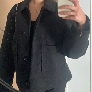 Coat/jacka - Stilren mörkgrå jacka från Lager 157 i ullblandning. Jackan har klassisk krage, stora framfickor och knäppning med knappar framtill. Passformen är loose och modellen är enkel men trendig, perfekt för lager på lager under höst och vinter.