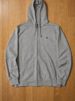 Grå zip hoodie från Polo Ralph Lauren - Klassisk grå hoodie med dragkedja från Polo Ralph Lauren. Hoodien har huva med dragsko, två fickor framtill och den ikoniska mörkblå loggan broderad på bröstet. Tillverkad i mjuk bomullsblandning för skön komfort och avslappnad stil.