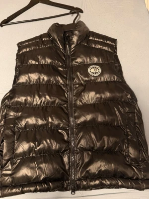  Canada Goose väst  - Snygg svart dunväst från Canada Goose med glansig finish och broderad logga på bröstet. Västen har hög krage, dragkedja framtill och två stora fickor. Perfekt för lager på lager och ger en cool streetwear-look.