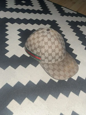 Gucci keps - Snygg keps från Gucci i beige med klassiskt GG-monogram över hela kepsen. På sidan finns de ikoniska gröna och röda ränderna. Kepsen har böjd skärm och är tillverkad i canvasmaterial. Perfekt för dig som vill sticka ut med en lyxig accessoar.