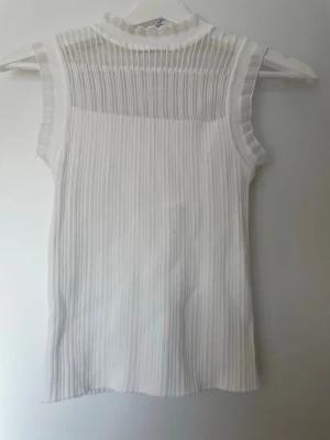 Vit ribbad ärmlös topp med meshdetalj - Säljer en vit ärmlös topp med ribbad struktur och transparent mesh upptill. Toppen har en liten volangkant vid halsen och materialet känns mjukt och stretchigt. Pris går att diskutera 💘