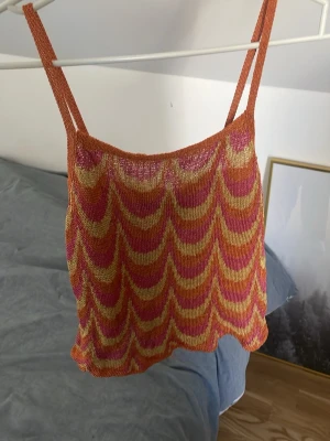 Mönstrad linnetopp - Säljer en färgglad stickad linnetopp med vågigt mönster i orange, rosa och gul. Toppen har smala axelband och är ärmlös, perfekt för varma dagar. Materialet är tunt och luftigt, vilket ger en skön känsla. Ingen synlig logga eller märke. Materialet är ganska hårt