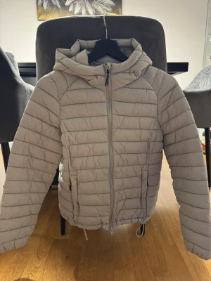 Ljusgrå pufferjacka med huva - Säljer en ljusblå/grön pufferjacka med huva och dragkedja framtill. Jackan har två sidofickor med dragkedja och justerbar dragsko i nederkant. Quiltad design med horisontella sömmar ger en clean och modern look. Perfekt för kalla vinterdagar.