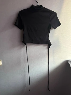 Topp - Snygg svart croppad topp med korta ärmar. Toppen har långa snören i sidorna och öppen rygg. Perfekt att styla med höga jeans eller kjol.