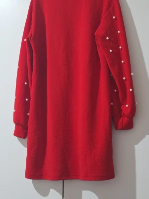 Röd klänning med pärldetaljer - Trendig röd klänning med långa ärmar dekorerade med vita pärlor. Klänningen har en rak passform och är gjord i ett mjukt syntetmaterial. Perfekt för dig som vill sticka ut med en unik detalj på ärmarna.