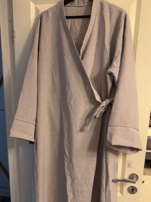 Ljusgrå långklänning med knytband - En stilren ljusgrå långklänning i mjukt tyg med långa ärmar och knytband i midjan. Klassisk kimono-inspirerad design med breda ärmslut och enkel krage. Perfekt för att chilla hemma och hålla sig bekväm. Köpt från Twmodest. Prislapp kvar. One size