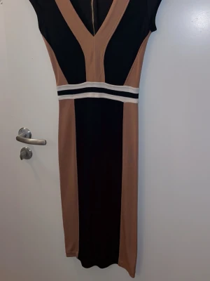 Svart och beige klänning med v-ringning - Snygg svart och beige klänning med markerad v-ringning och breda kontrasterande paneler. Klänningen har en vit och svart randig detalj i midjan och är ärmlös. Perfekt för dig som gillar stilrena och moderna plagg.