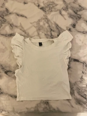 Vit ribbad topp med volangärmar SHEIN - Säljer en söt vit ribbad topp från SHEIN i storlek M. Toppen har korta volangärmar och rund halsringning. Materialet är stretchigt och mjukt, perfekt för en trendig och bekväm look. Passar till jeans eller kjol för en fräsch outfit.