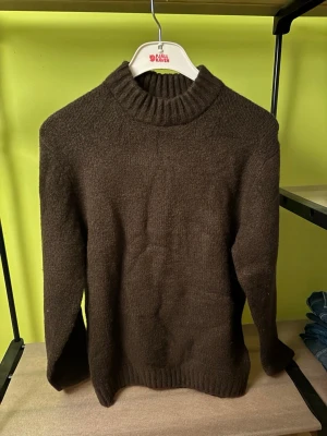 Mörkbrun stickad tröja från Rebel - Mysig mörkbrun stickad tröja från Rebel med hög ribbad krage och ribbade muddar vid ärmslut och nederkant. Enkel och stilren design som passar perfekt till höstens och vinterns outfits. Tillverkad i mjukt material för extra komfort.