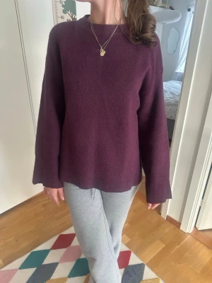 Mörklila oversized stickad tröja - Mysig oversized stickad tröja i mörklila färg med rund halsringning och långa, vida ärmar. Tröjan har en rak passform och ribbstickade muddar vid ärmslut och nederkant. Perfekt för lager-på-lager och chill vardagsstil.