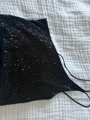 Svart paljett tubtopp med smala band - Snygg svart tubtopp med tunna axelband och glittriga paljetter över hela toppen. Perfekt för dig som vill sticka ut och glänsa lite extra. Toppen har en croppad passform och är tillverkad i ett stretchigt syntetmaterial.