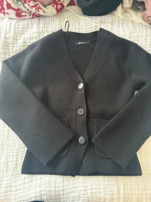 Svart kofta från Gina Tricot - Mysig svart kofta från Gina Tricot med tre stora knappar framtill och två fickor. Koftan har v-ringning och långa ärmar, perfekt att slänga på sig över en t-shirt. Materialet är mjukt och stickat.