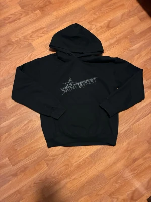 Svart Saint Laurent hoodie med tryck - Svart hoodie från Saint Laurent med stor huva och magficka. Framtill finns ett coolt tryck med stjärna och Saint Laurent-logga i grått. Klassisk passform och mjukt material, perfekt för en avslappnad look len och mjuk på insidan, bra skick nästan ny! 