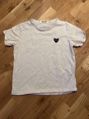Vit t-shirt från Comme des Garçons Play - Vit t-shirt från Comme des Garçons Play med det ikoniska svarta hjärtat med ögon broderat på bröstet. Klassisk passform och rund halsringning. Perfekt basic-plagg med en twist för dig som gillar streetwear och stilrena detaljer.
