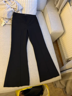 Svarta utsvängda kostymbyxor Gina Tricot - Säljer ett par svarta kostymbyxor från Gina Tricot med utsvängda ben och hög midja. Byxorna har en klassisk look och är sydda i ett mjukt, följsamt tyg som ger en snygg siluett. Perfekta för dig som gillar stilrena och trendiga plagg. Storlek M
