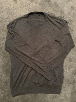 Mörkgrå stickad tröja från Riley - Snygg mörkgrå stickad tröja från Riley med rund halsringning. Tröjan är tunnstickad och har ribbade muddar vid ärmslut och nederkant. Perfekt för lager-på-lager och enkel att matcha med olika outfits.