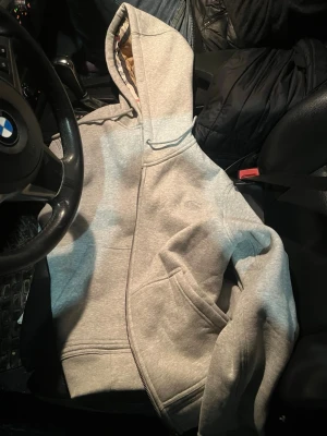Grå hoodie från Burberry - Säljer en stilren grå hoodie från Burberry med dragkedja och klassisk huva. Hoodien har fickor framtill och diskret Burberry-logga broderad på bröstet. Perfekt för dig som gillar enkel men exklusiv streetwear. Materialet är mjukt och skönt bomullstyg.