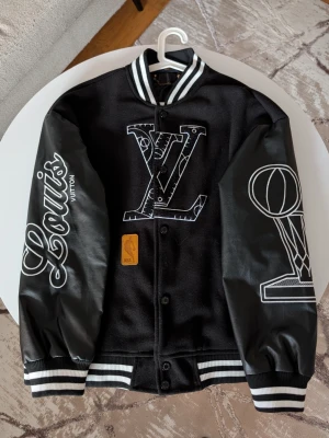 Iouis Vuitton jacka NBA edition  - Denna stilrena baseballjacka kombinerar en klassisk sportestetik med lyxiga detaljer. Jackan är designad i en tidlös färgskala av svart och vitt, vilket skapar en skarp och modern kontrast.