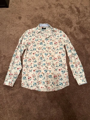 Blommig skjorta från Lindbergh - Snygg vit skjorta från Lindbergh med blommigt mönster i blått och rött. Skjortan har lång ärm, klassisk krage och knappar framtill. Tillverkad i bomull och har en modern fit som sitter riktigt bra. Storlek M