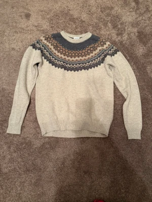 Beige stickad tröja från Kappahl - Stickad tröja från Kappahl i beige med rund hals och snyggt mönster i brunt, mörkgrått och vitt över bröstet och axlarna. Tröjan har ribbade muddar vid ärmslut och nederkant. Perfekt för kalla dagar och riktigt mysig stil. Storlek M