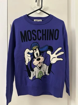 Lila stickad tröja H&M x Moschino - Säljer en lila stickad tröja från H&M x Moschino med stor Goofy-figur framtill och svart MOSCHINO-tryck. Tröjan har rund halsringning och långa ärmar. Perfekt för dig som gillar statement-plagg och Disney!