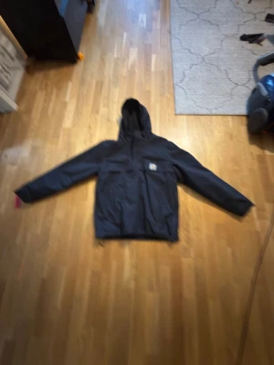 Svart Carhartt pullover - Snygg svart vindjacka från Carhartt med huva och dragkedja. Jackan har två stora fickor framtill och en liten Carhartt-logga på bröstet. Perfekt för blåsiga dagar och enkel att matcha med olika outfits.  Använd 2-3 gånger