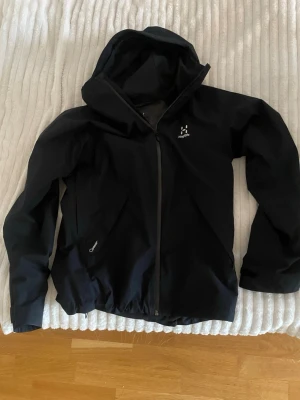Svart vindjacka från Haglöfs - Snygg svart vindjacka från Haglöfs med huva och hel dragkedja framtill. Jackan har två sidofickor med dragkedja och diskret logga på bröstet. Tillverkad i ett lätt och slitstarkt material som står emot vind. Perfekt för utomhusaktiviteter och streetwear. Priset kan diskuteras 