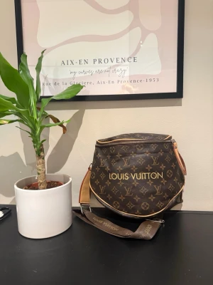  brun axelväska - Säljer en brun axelväska  med klassiskt monogrammönster i guld och beige detaljer. Väskan har justerbar axelrem med logga, dragkedja upptill och rundad form. Perfekt för dig som vill ha en ikonisk och trendig accessoar.