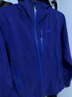 Blå vindjacka från Patagonia - Snygg blå vindjacka från Patagonia med huva och vattentäta dragkedjor. Jackan har tre fickor med dragkedja och är tillverkad i ett lätt och slitstarkt material. Perfekt för blåsiga dagar och outdoor-äventyr.