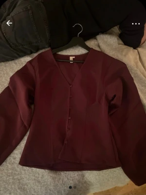 Vinröd blus med knappar från Nelly - vinröd blus från Nelly med v-ringning och knappar framtill. Blusen har lång ärm och är tillverkad i ett tunt, lätt mattetal. Jättefint skick! 🩷