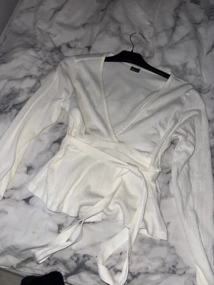 Vit omlottopp med knytband från Gina Tricot - Säljer en stilren vit omlottopp med långa ärmar och knytband i midjan. Toppen har v-ringad hals och är tillverkad i ett lätt, strukturerat tyg som ger en snygg passform. Perfekt att styla med jeans eller kjol för en trendig look.