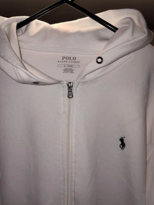 Vit hoodie från Polo Ralph Lauren - Snygg vit hoodie med dragkedja från Polo Ralph Lauren. Klassisk design med huva, fickor och den ikoniska broderade loggan på bröstet. Tillverkad i mjukt bomullsmaterial som känns skönt mot huden. Perfekt för en clean och stilren look.