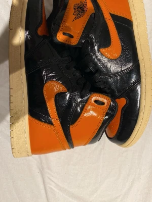 Nike Air Jordan 1 Shattered Backboard - Säljer ett par Nike Air Jordan 1 Shattered Backboard med svart och orange patentläder och beige sula. Klassisk high-top siluett med ikonisk vinge-logga vid ankeln och svart snörning. Perfekta för dig som vill sticka ut med en fet färgkombo.och det kommer inte sko kartong