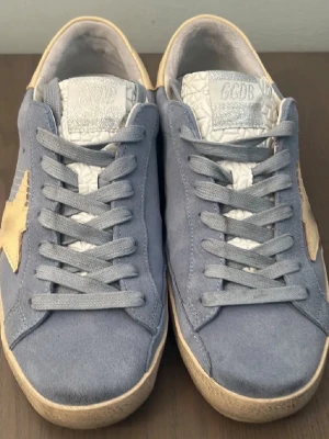 Blåa Golden Goose sneakers med stjärna - Snygga sneakers från Golden Goose i blå mocka med grå skosnören och beige stjärndetalj på sidan. Skorna har en låg siluett och vit texturerad plös med märkets logga. Yttersulan är beige och ger en cool kontrast till den blå färgen.