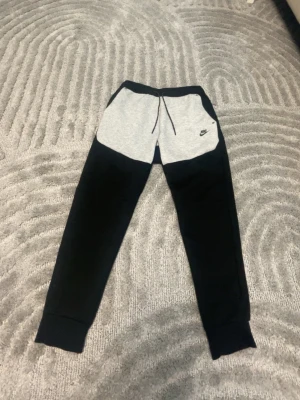 Nike mjukisbyxor svart/grå - Snygga mjukisbyxor från Nike i svart och ljusgrått med klassisk logga på vänster sida. Byxorna har dragsko i midjan, muddar vid bensluten och är tillverkade i mjukt sweatshirtmaterial. Perfekta för chill eller träning.