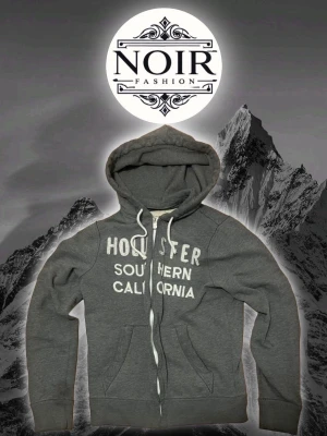 Grå hoodie från Hollister - Mörkgrå hoodie från Hollister med vit dragkedja och tryck 'Southern California' på bröstet. Klassisk huva med dragsko och två fickor framtill. Perfekt för en avslappnad och cool stil.